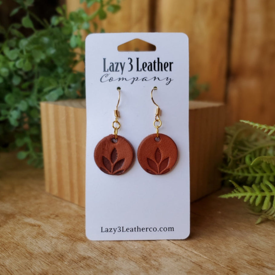 Small round chestnut-colored leather earrings with gold hooks and a hand-stamped leaf design, made by Lazy 3 Leather Co.