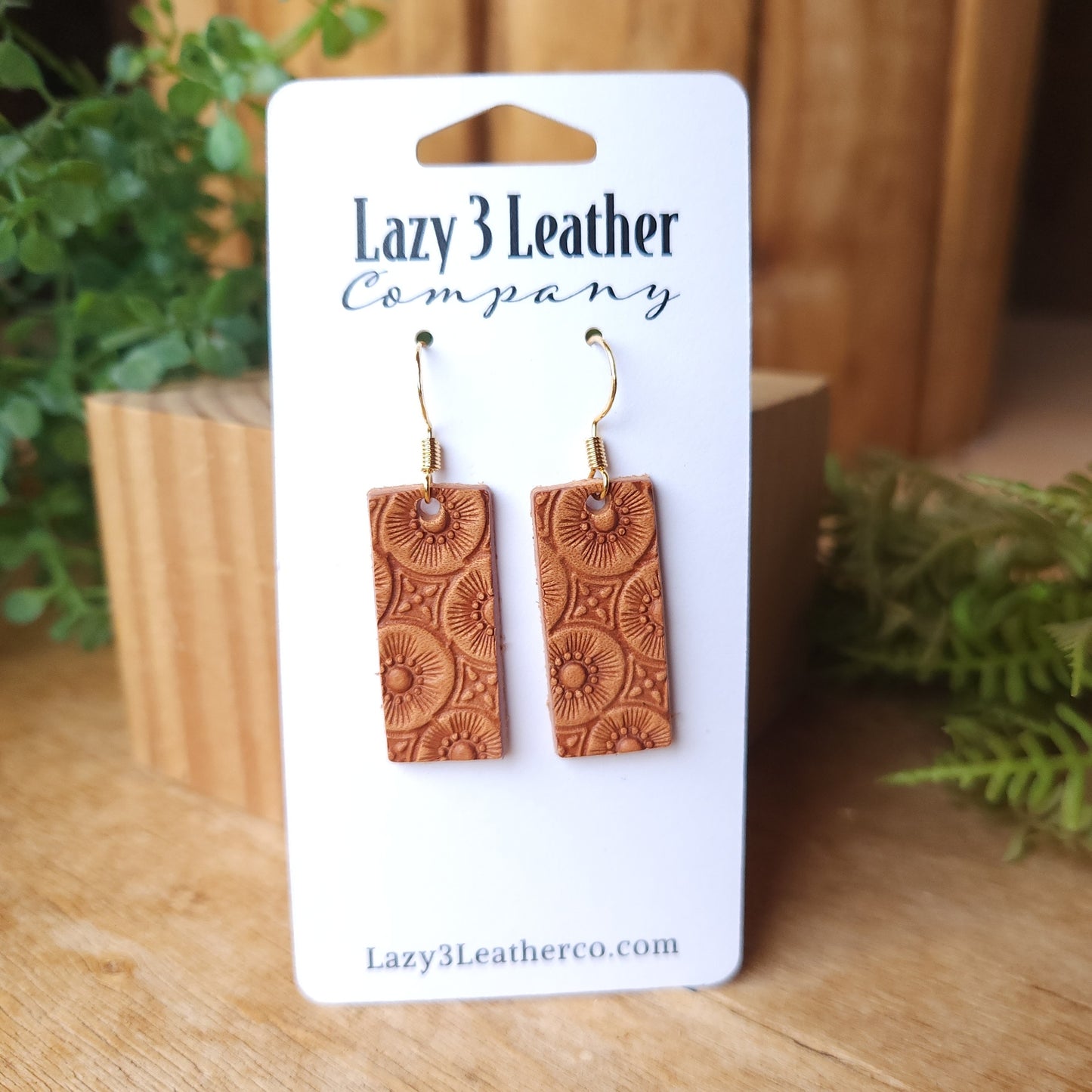 Handmade leather rectangle earrings stamped with a sunburst design, displayed on a Lazy 3 Leather Company card with a rustic wood and greenery background.