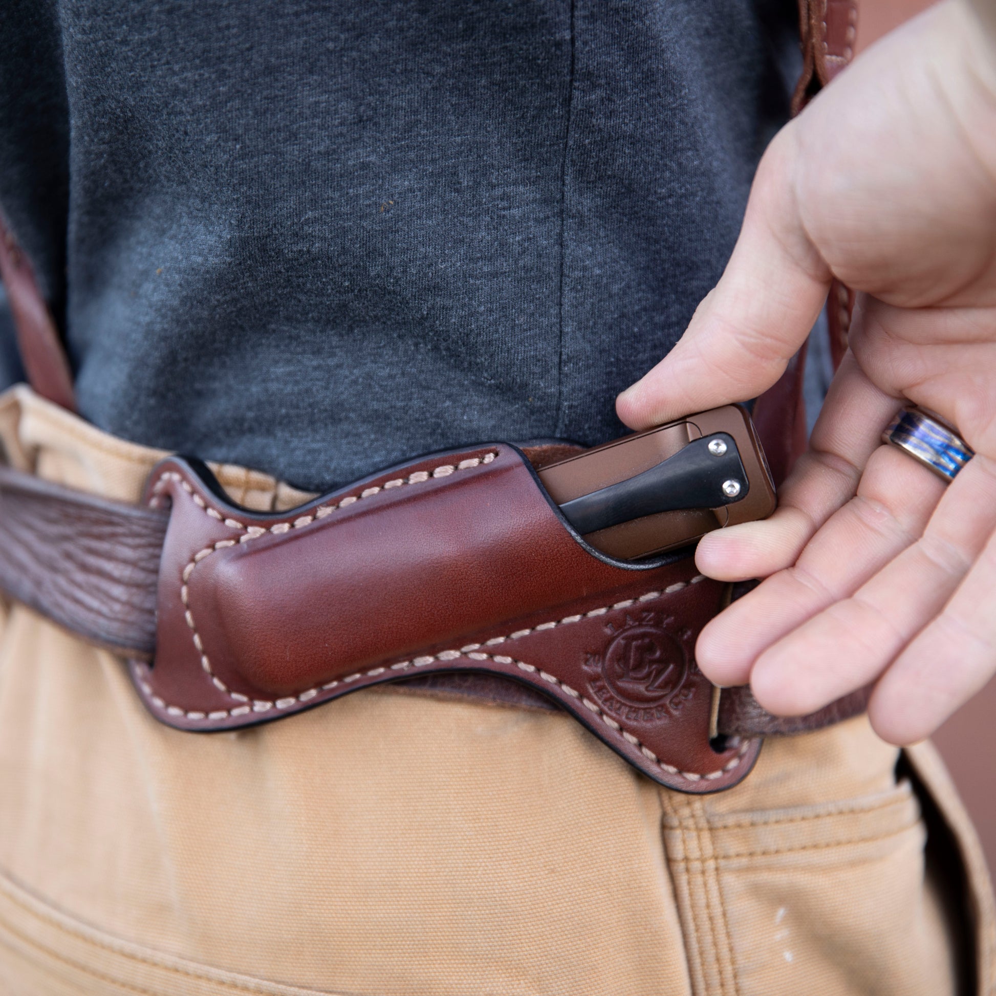 Person holding a brown leather knife sheath with a Arkfelt Flashlight inside, wearing a gray shirt and beige pants.