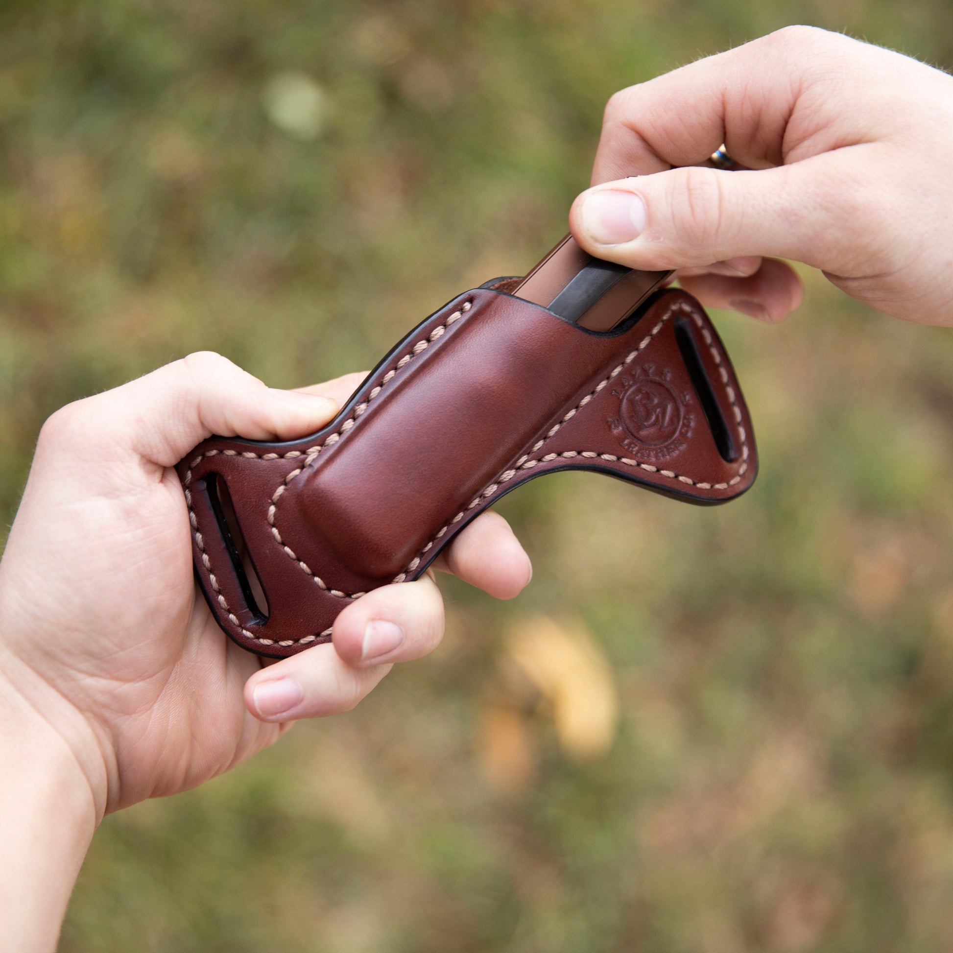 Brown leather sheath with Arkfelt flashlight being held by two hands against a blurred natural background
