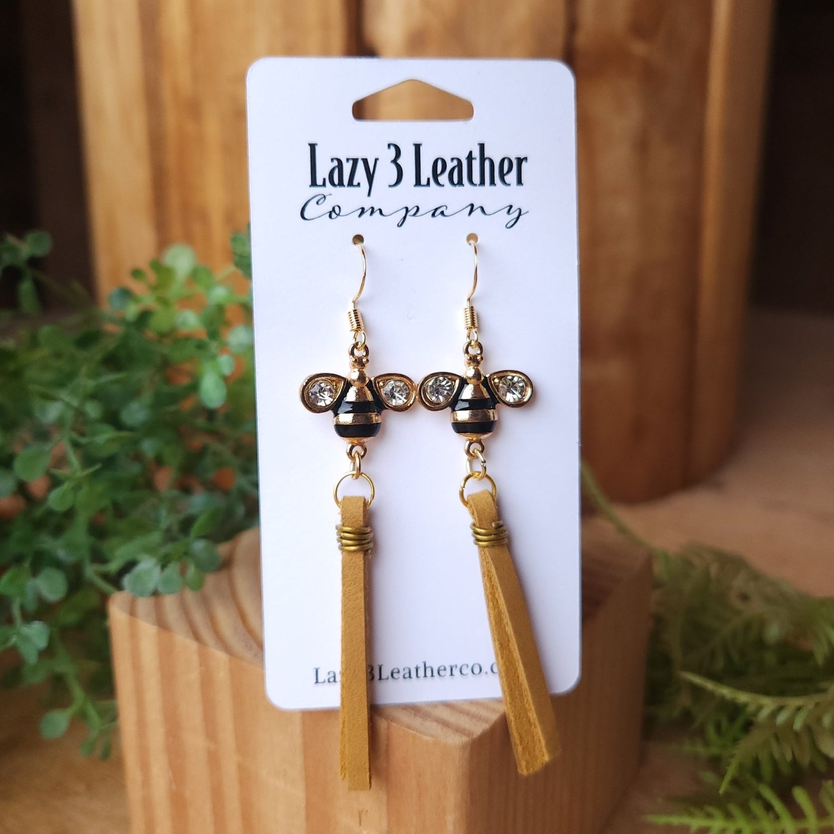 Handmade earrings featuring gold-tone bee charms with rhinestone wings and mustard-yellow leather tassels, displayed on a Lazy 3 Leather Company card with a rustic wood and greenery background.