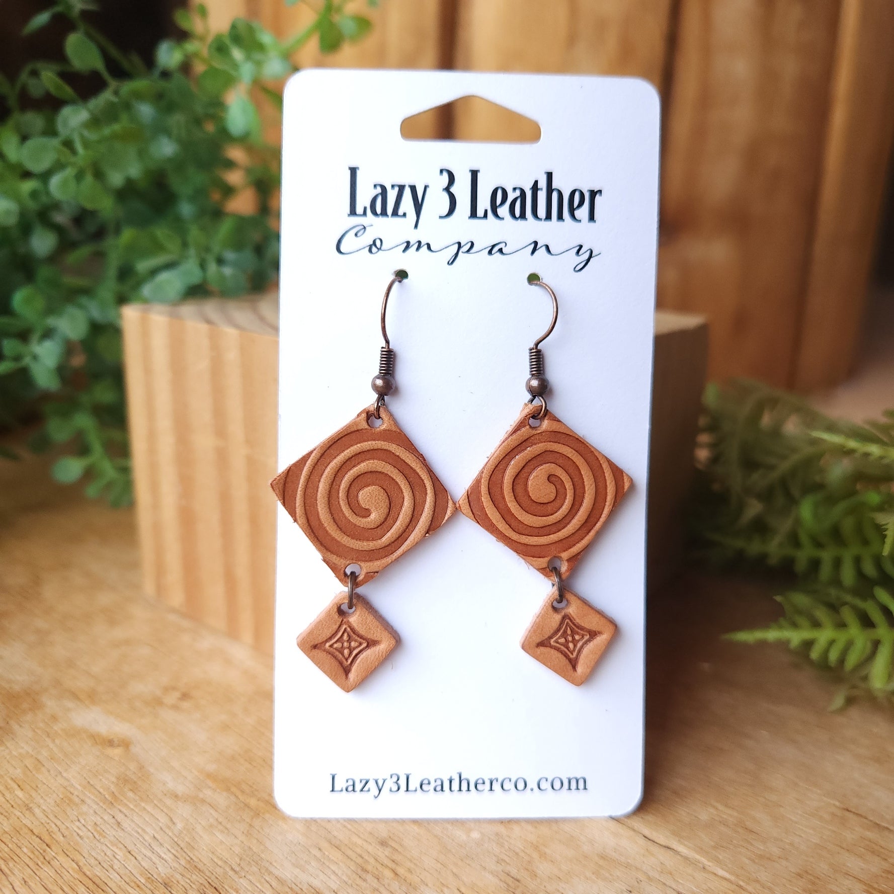Handmade leather earrings with a stamped swirl design and small diamond dangle, displayed on a Lazy 3 Leather Company card with a rustic wood and greenery background.