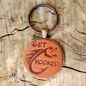 Hand stamped leather keychain that says Get Hooked with a fish stamped into the leather. This keychain has been mounted in a round metal pendant with a 1 inch antique copper keyring. This keychain is made using veg tanned Wickett & Craig leather.
