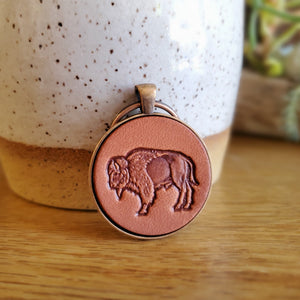 Hand stamped leather keychain with a buffalo on it that has been mounted in a round metal pendant with a 1 inch antique copper keyring. This keychain is made using veg tanned Wickett & Craig leather.